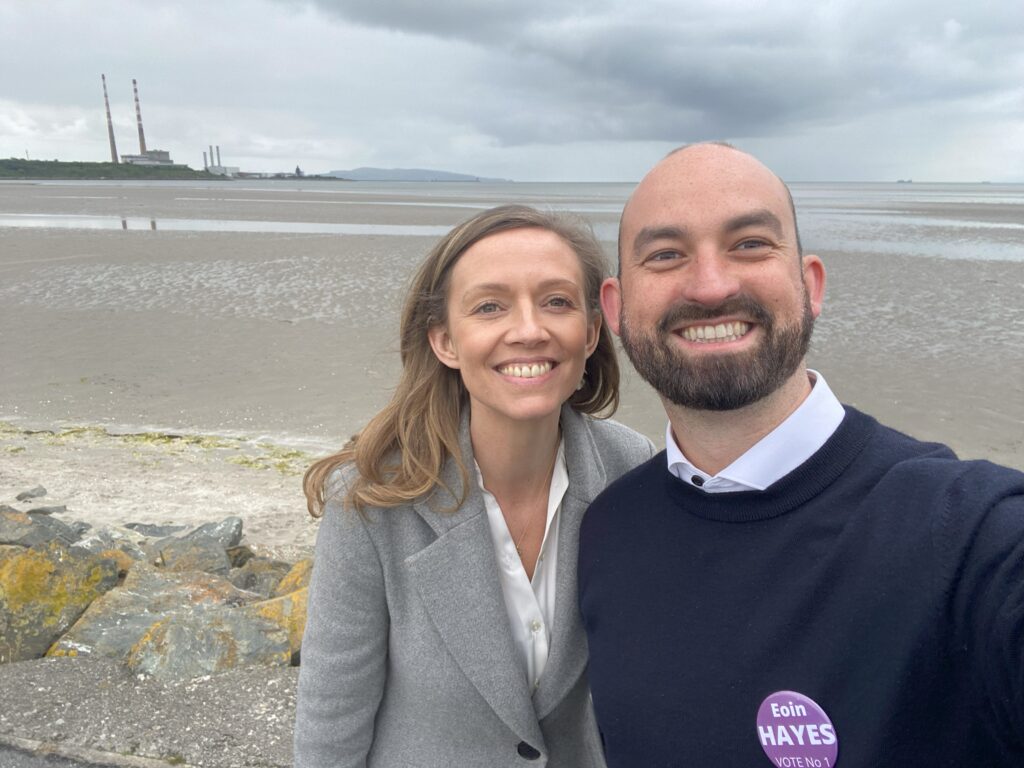 A smiley selfie of Holly Cairns and Eoin Hayes on Sandymount beach, with the Poolbeg chimney stacks visible in the background on a typically Irish cloudy day.