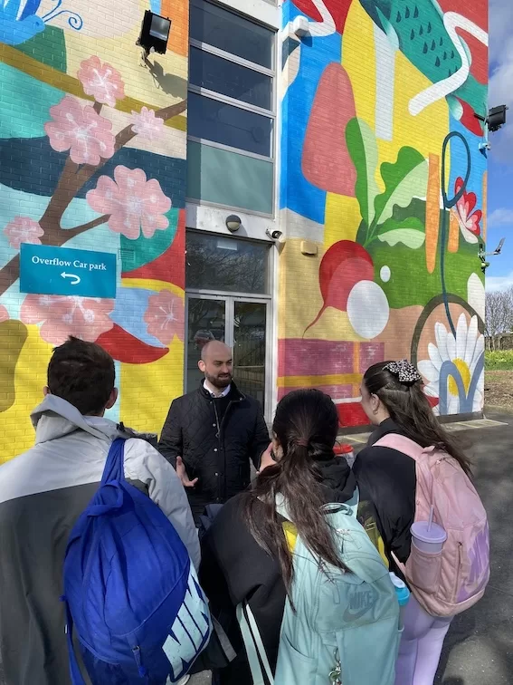 Eoin in front of a brightly painted school, talking to 3 teenage children with their backs to the camera.