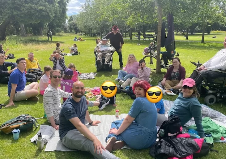 Eoin and his constituents and campaign team in a park, with folks sitting on picnic blankets and in mobility chairs, and kids with emojis over their faces for privacy purposes in the photo.