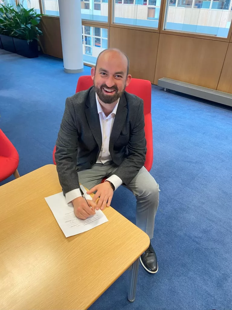 Eoin sitting to sign his documentation to officially join Dublin City Council, grinning joyously at the camera.