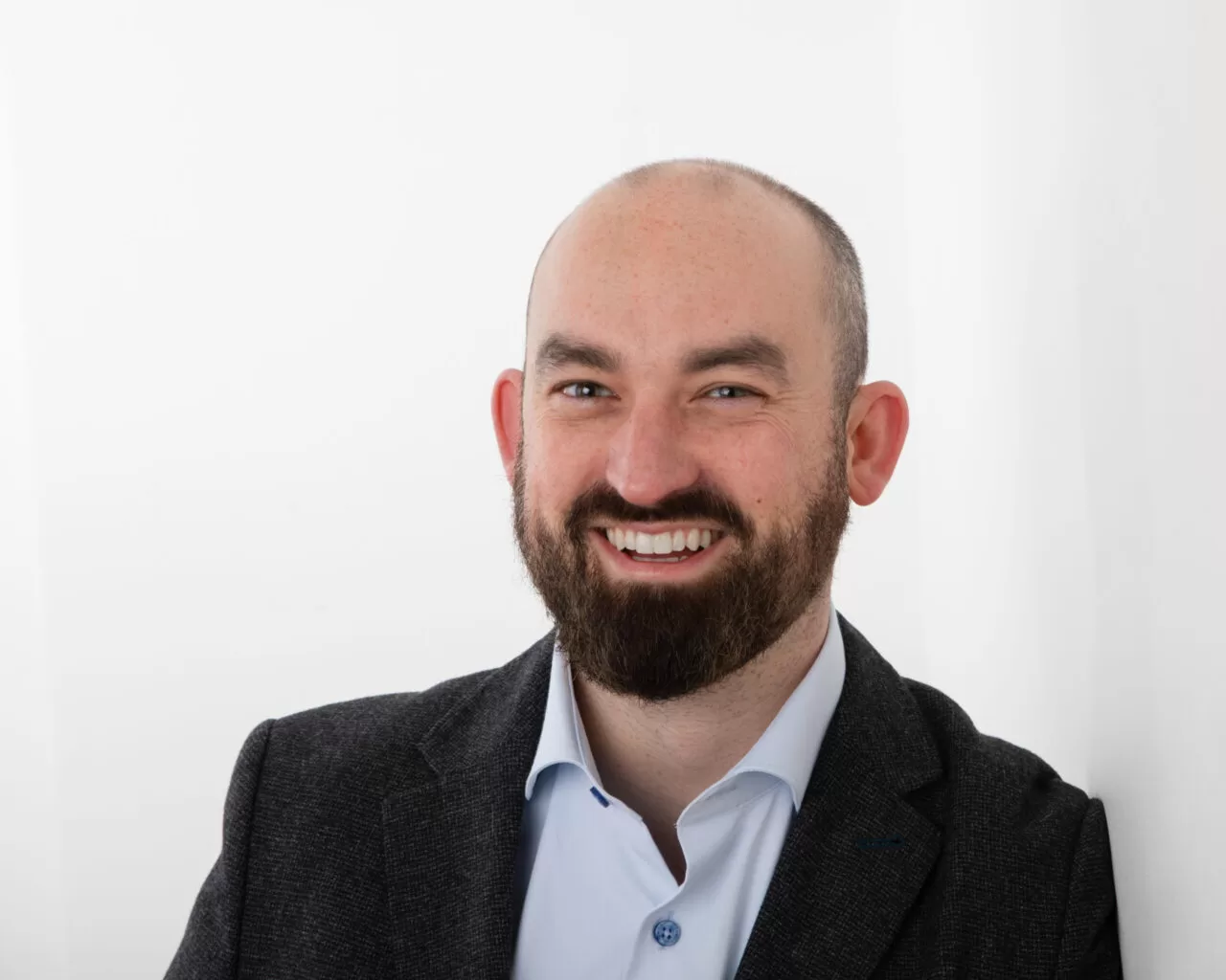 Eoin Hayes' professional headshot, where he is leaning against a wall in an open-collared shirt and jacket, smiling at the camera.