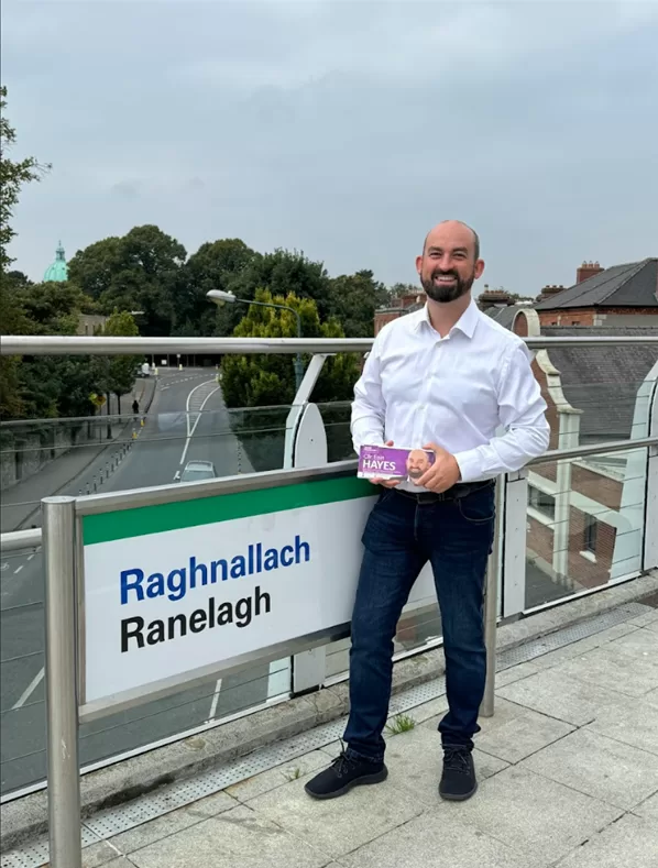 Eoin holding his campaign leaflets and grinning at the camera beside the Ranelagh Luas stop sign.